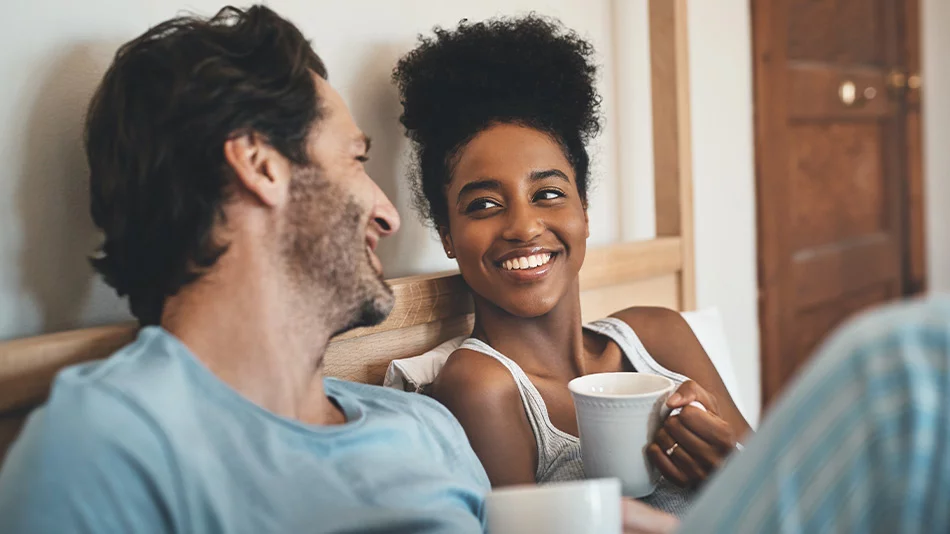 A young couple with a cup of coffee, smiling at each other on the bed in the morning.