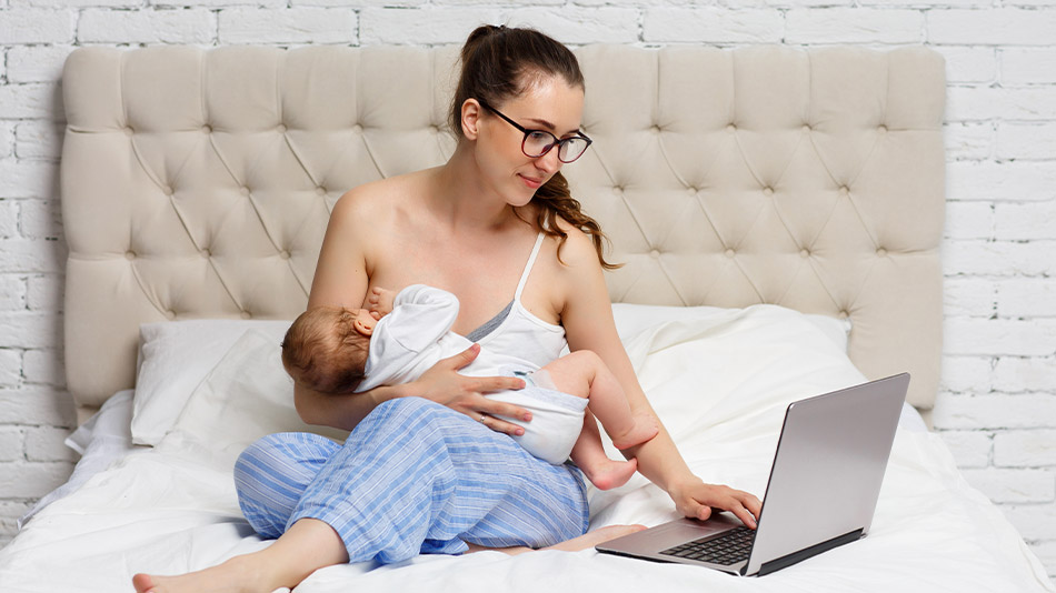 A woman with glasses breastfeeding her newborn while using her laptop.