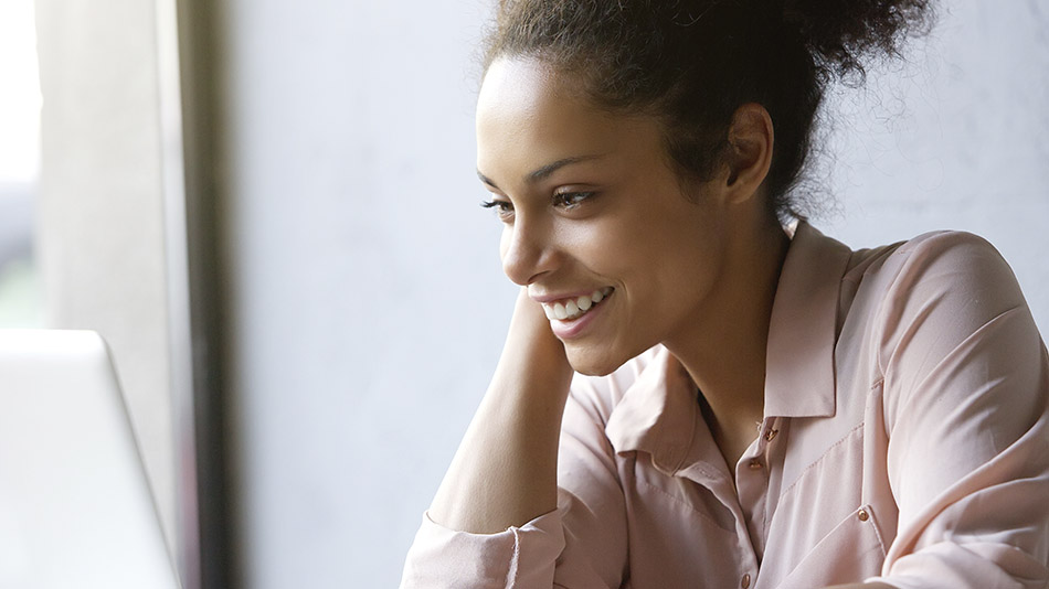 A cheerful young woman smiles while using her laptop.